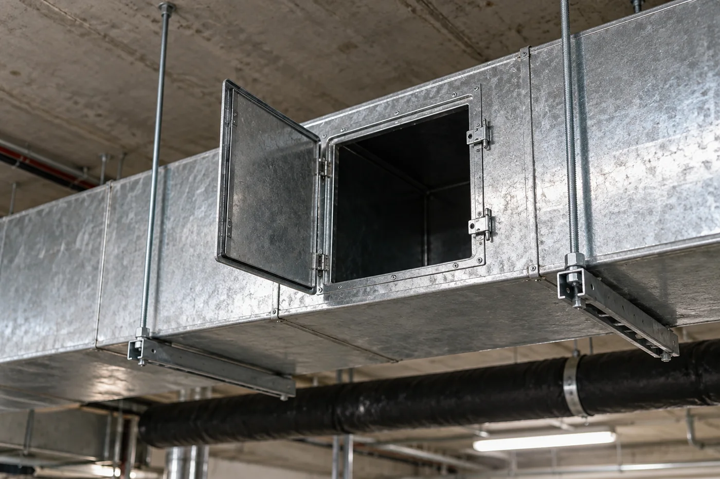 An open access panel on a galvanized duct beside hanger rods and adjacent pipework